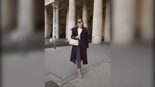 Woman in a long burgundy coat, white top, grey jeans and beige boots with a cream bag