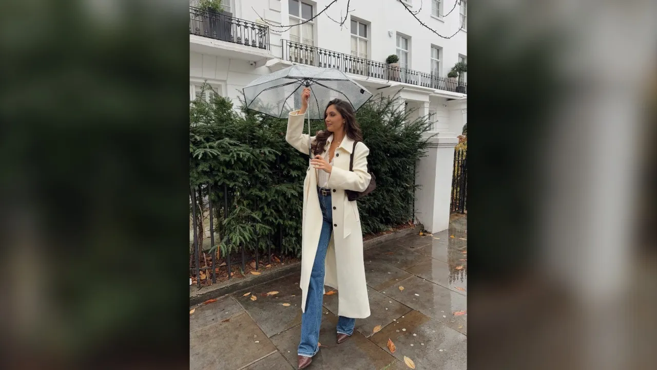 Woman in a long cream coat, brown top and blue jeans walking on a rainy street