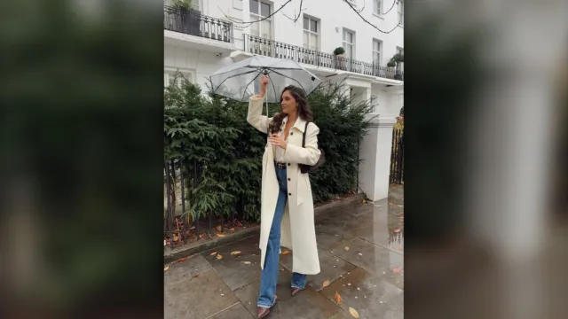 Woman in a long cream coat, brown top and blue jeans walking on a rainy street