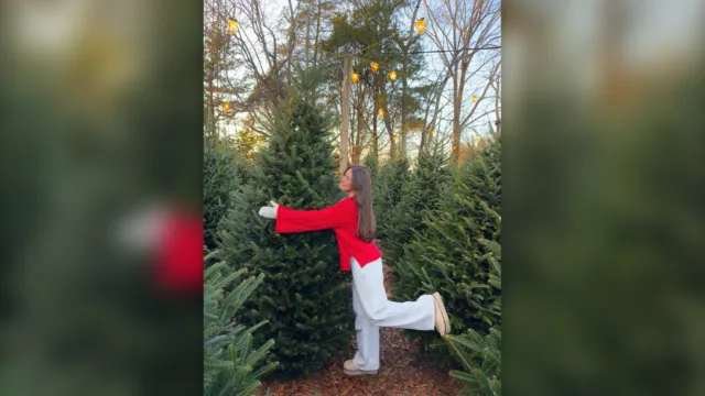 Woman in a red jacket and white jeans posing between Christmas trees at a tree farm