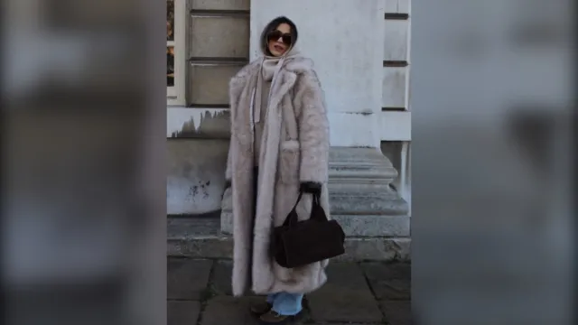 Woman in a long pale faux fur coat with a scarf over her head, jeans and a black bag