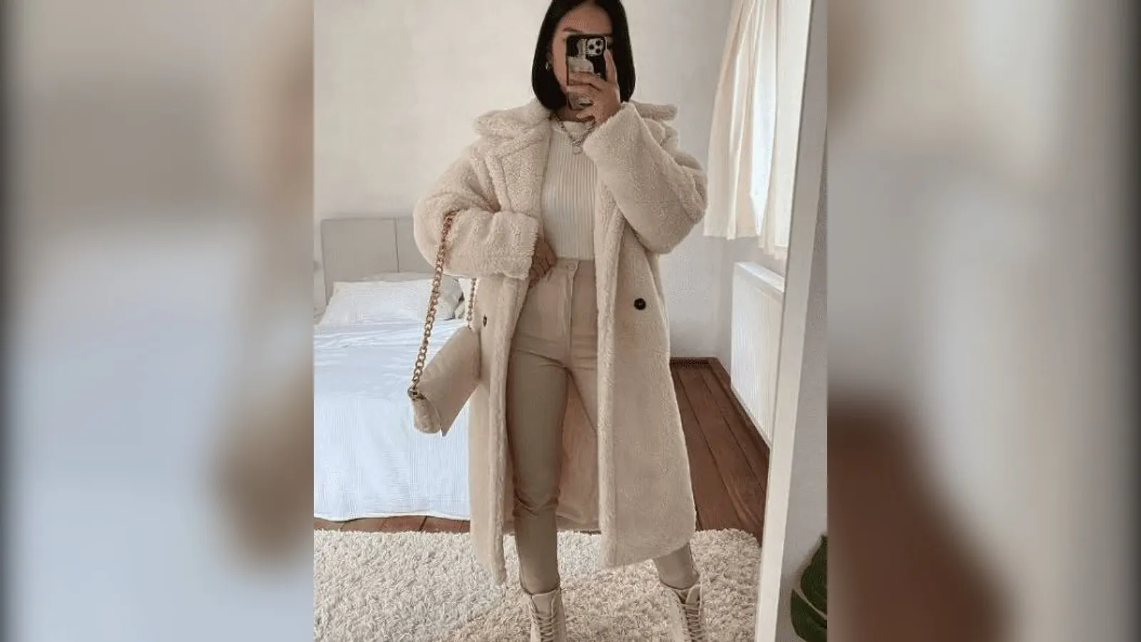 Woman in a plush long cream coat, creamy knit and beige pants taking a mirror selfie