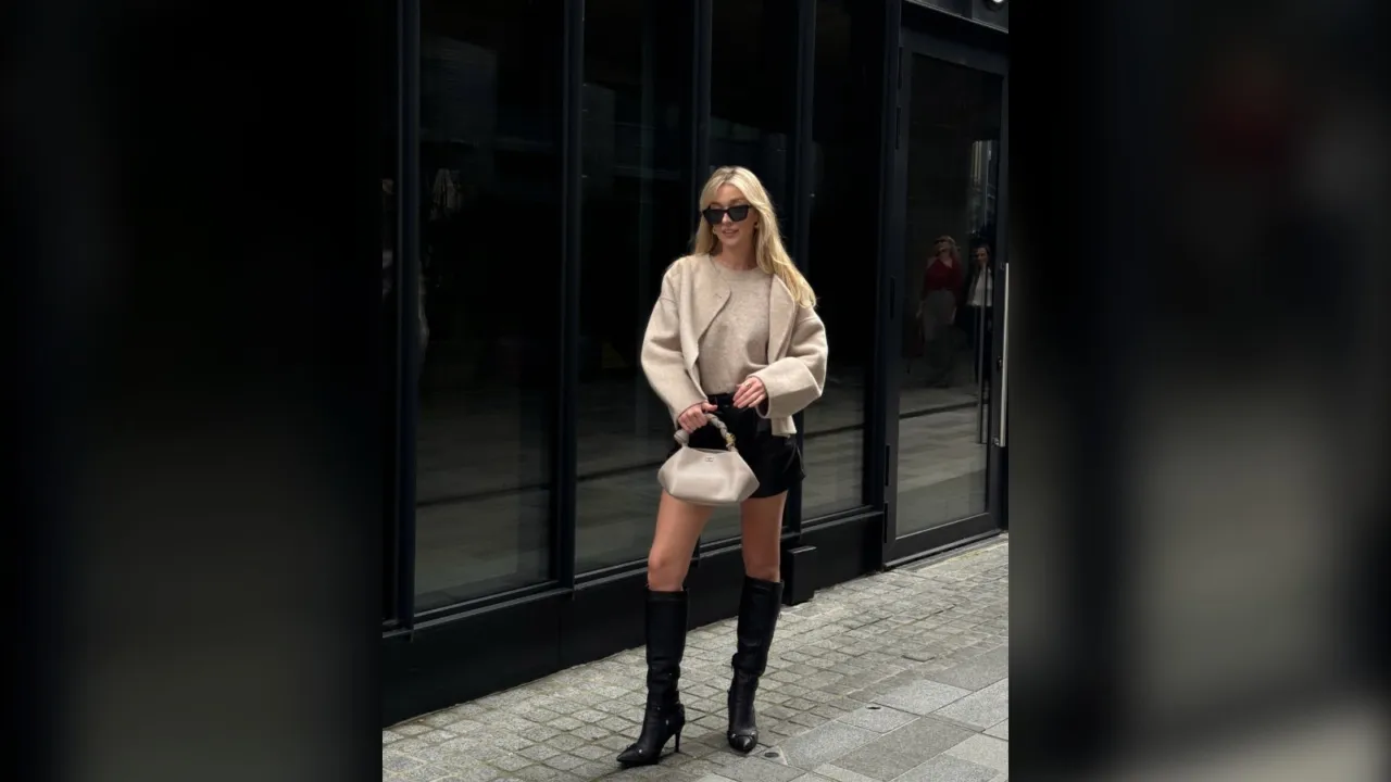 Woman in a cream sweater, black shorts and black knee high boots holding a cream bag