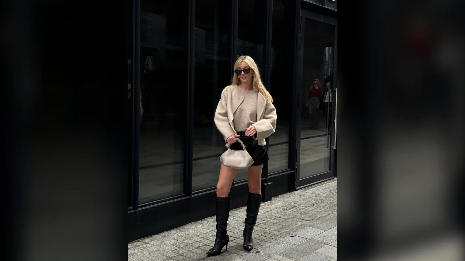 Woman in a cream sweater, black shorts and black knee high boots holding a cream bag