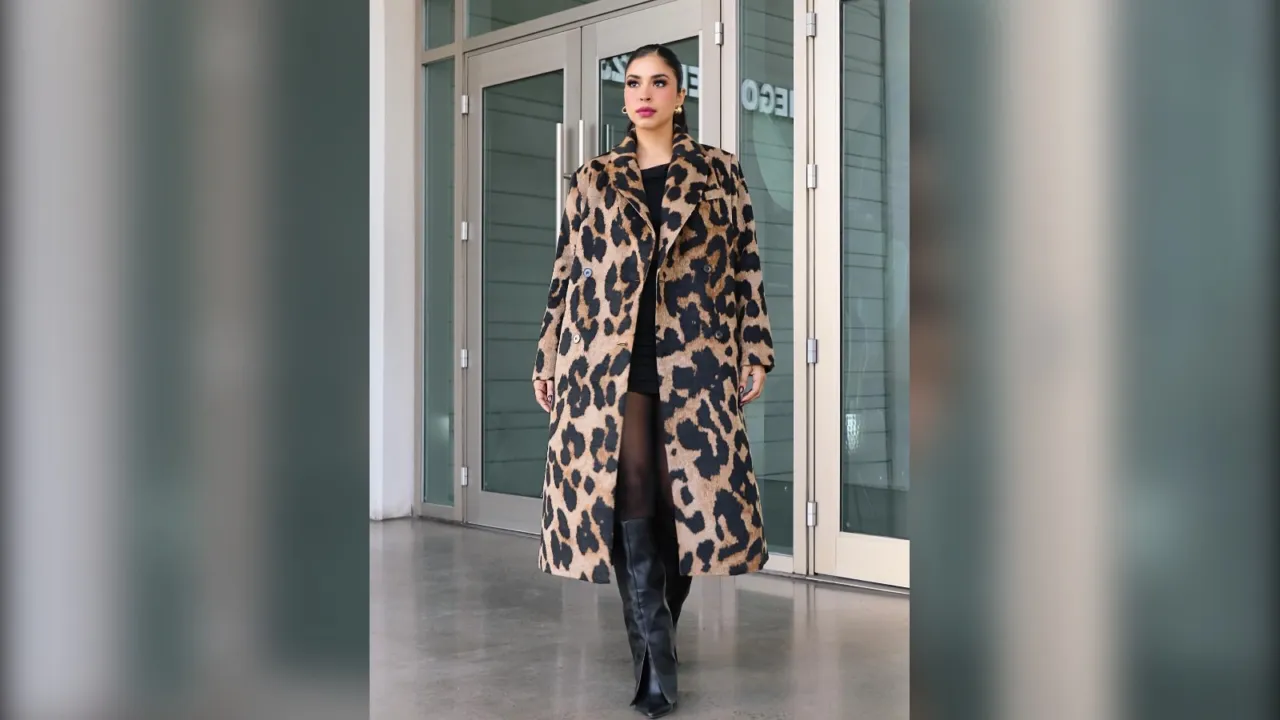 Woman in a long leopard print coat, sheer black tights and black knee high boots in front of glass doors