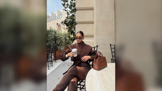 Woman in a brown knit set and long coat sitting outside with a coffee