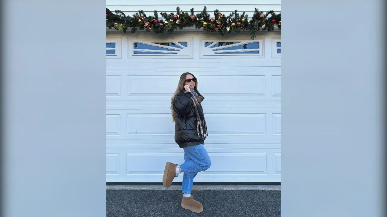 Woman in a black puffer jacket, light jeans and chestnut Ugg platform boots in front of a white garage door