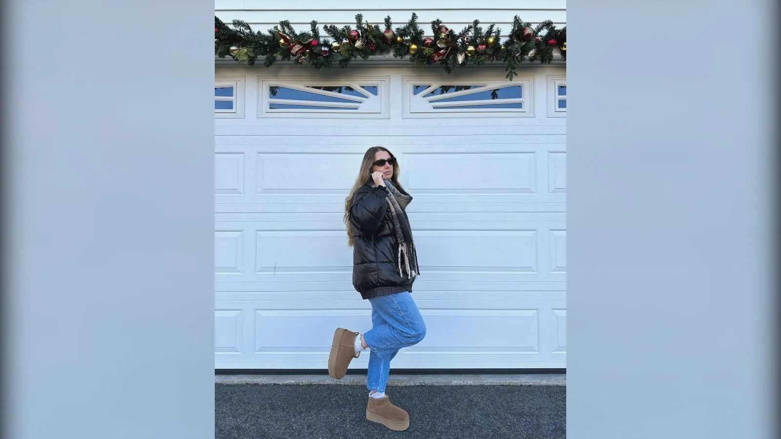 Woman in a black puffer jacket, light jeans and chestnut Ugg platform boots in front of a white garage door