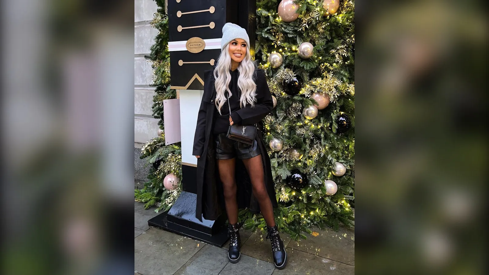 Woman in a long black coat, leather shorts, tights and a white beanie by a Christmas tree, winter concert outfit