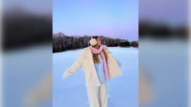Woman in a cream teddy coat, pink scarf and earmuffs in the snow, winter vacation outfit