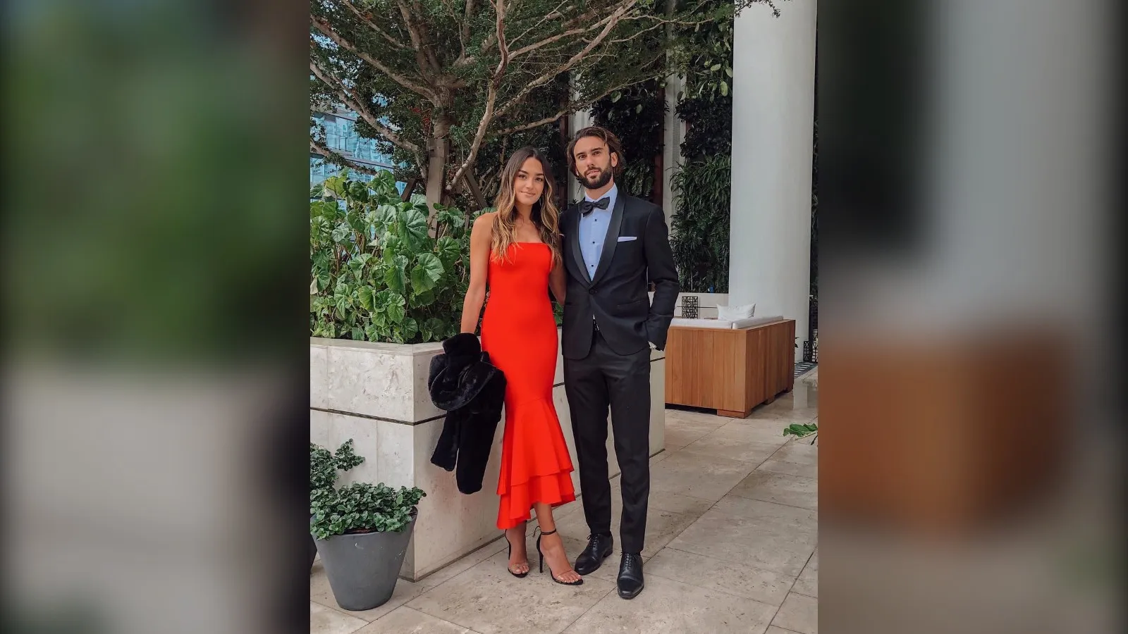 Wedding guest in a red mermaid dress holding a faux fur jacket next to her partner in a tuxedo